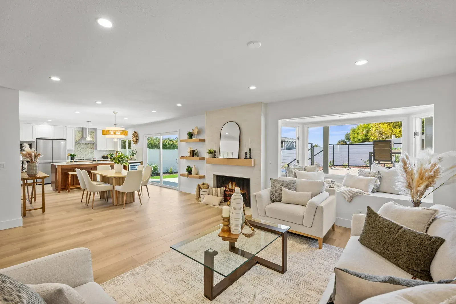Bright open-plan living room and kitchen with light wood floors, white furniture, and large windows overlooking a city skyline
