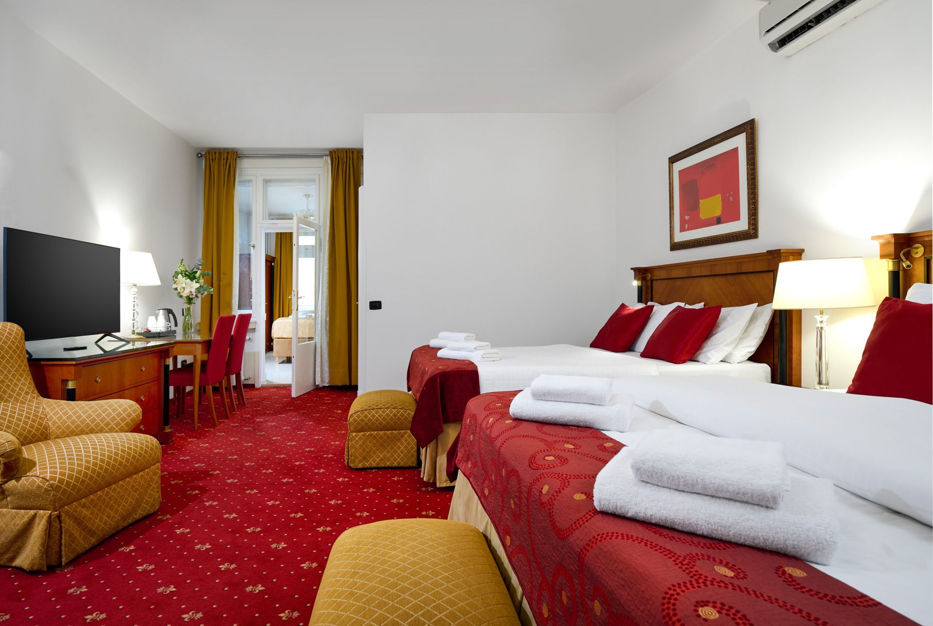 Hotel room with two beds, a desk, and a connecting room with another bed. Red carpet.