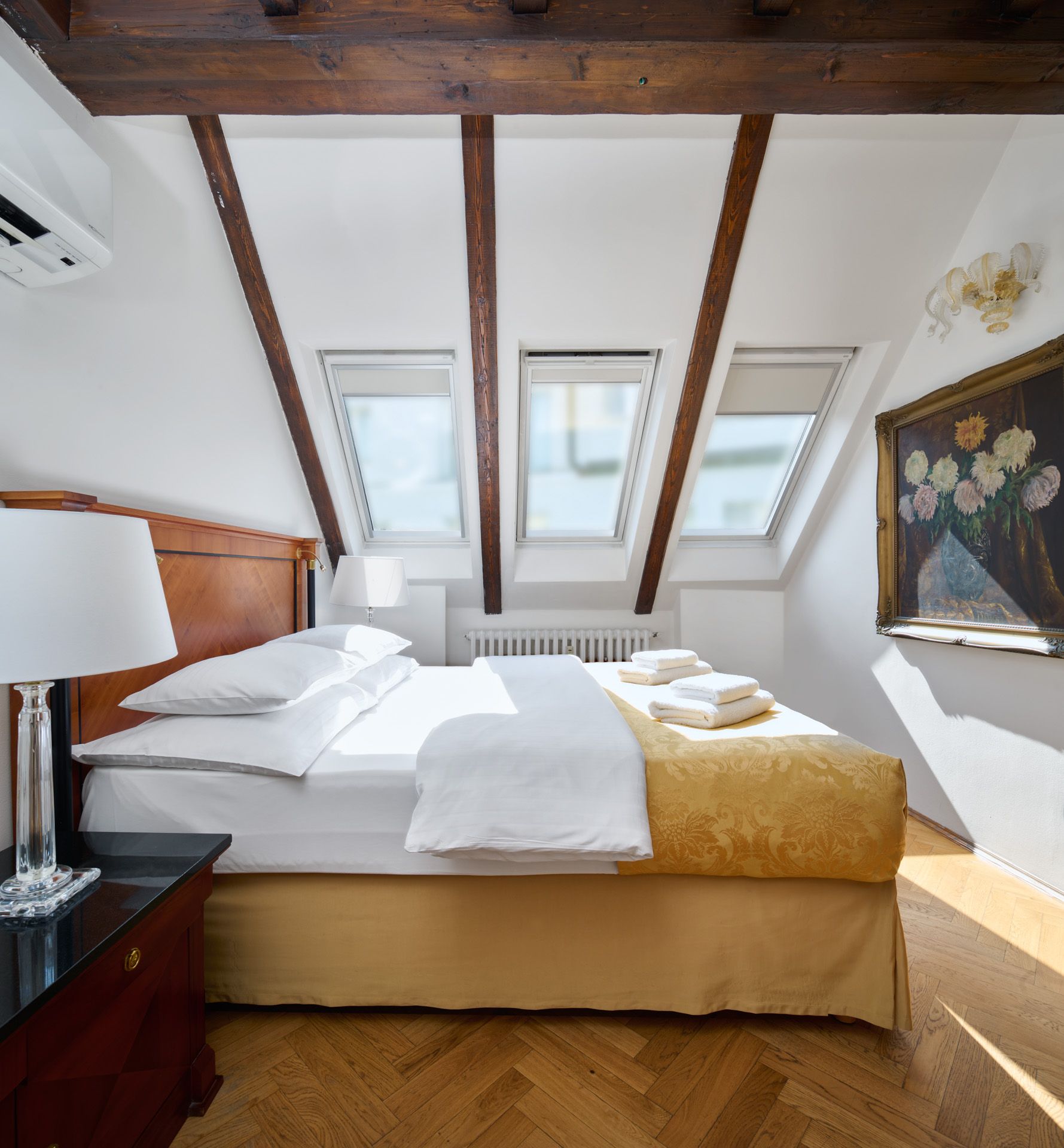 A hotel room with three beds, a desk, a TV, and an armchair. The room is white with gold and dark blue accents.