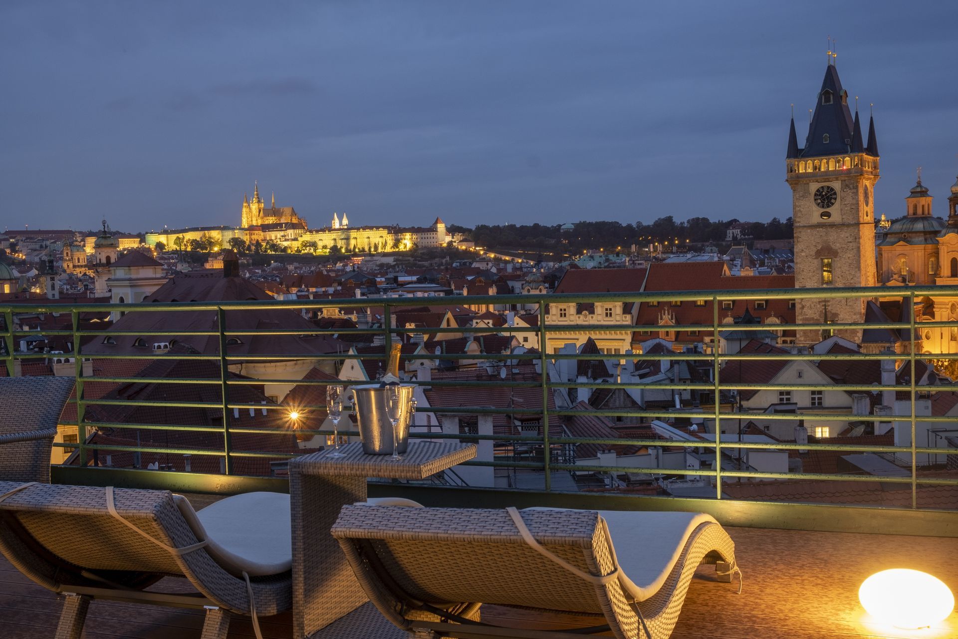Prague cityscape at sunset, featuring a river with boats, a bridge, and buildings with golden hues.