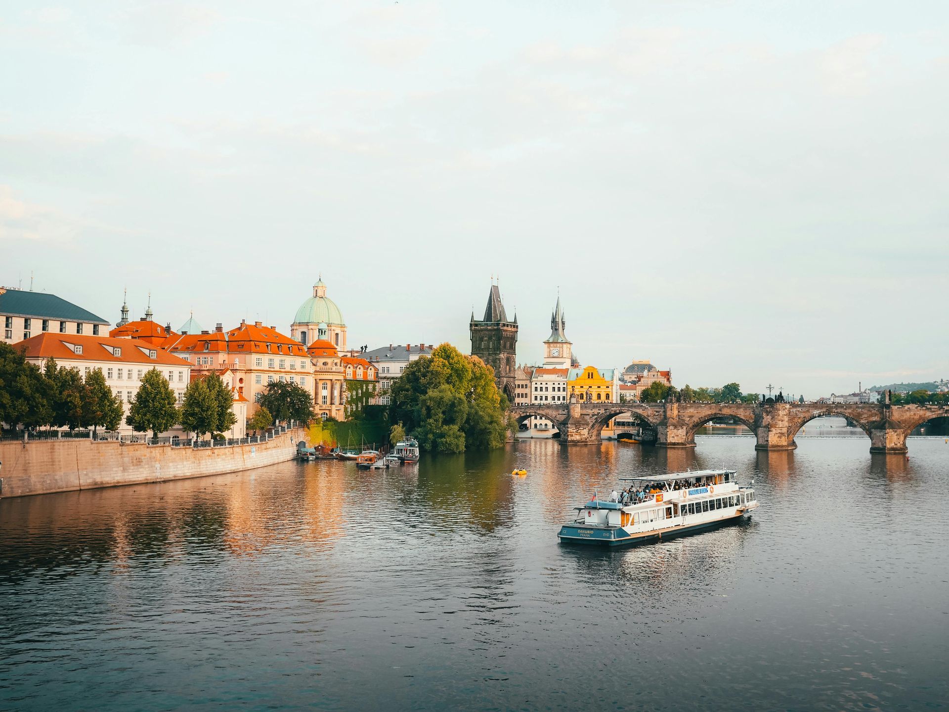 Prague cityscape with Charles Bridge, boat on Vltava River, buildings with red roofs under overcast sky.