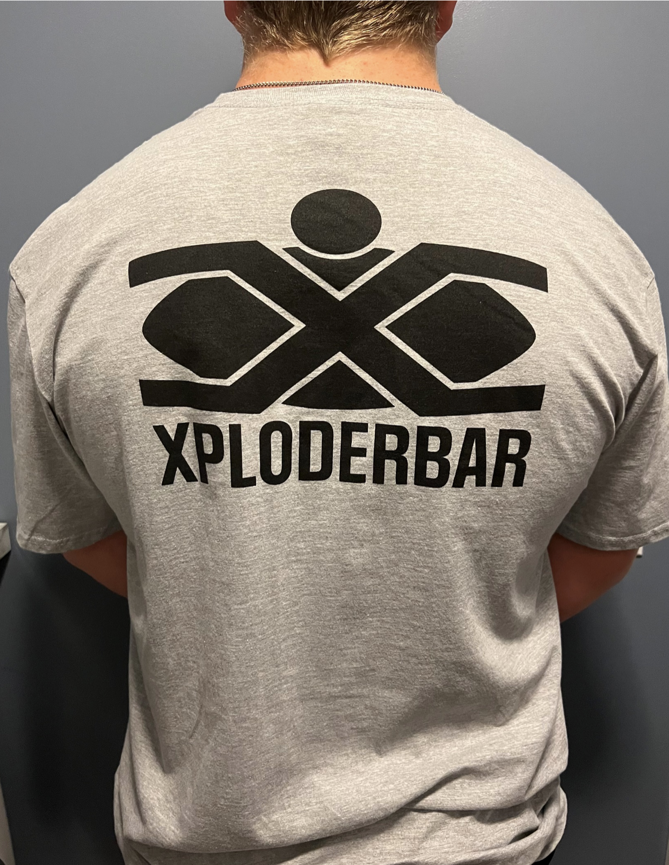 A man is wearing a grey t-shirt that says xploderbar on the back
