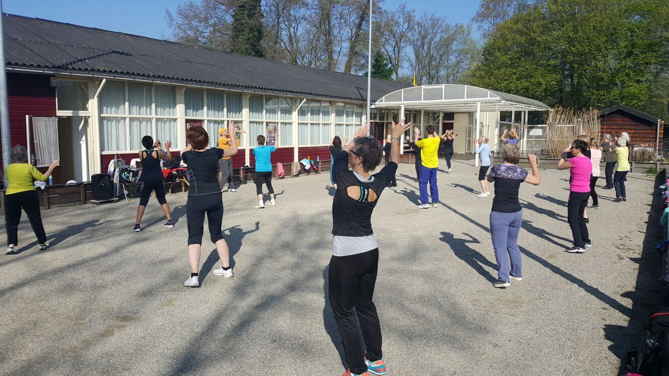 Een groep mensen sport buiten voor een gebouw, met hun armen omhoog. Zonnige dag.