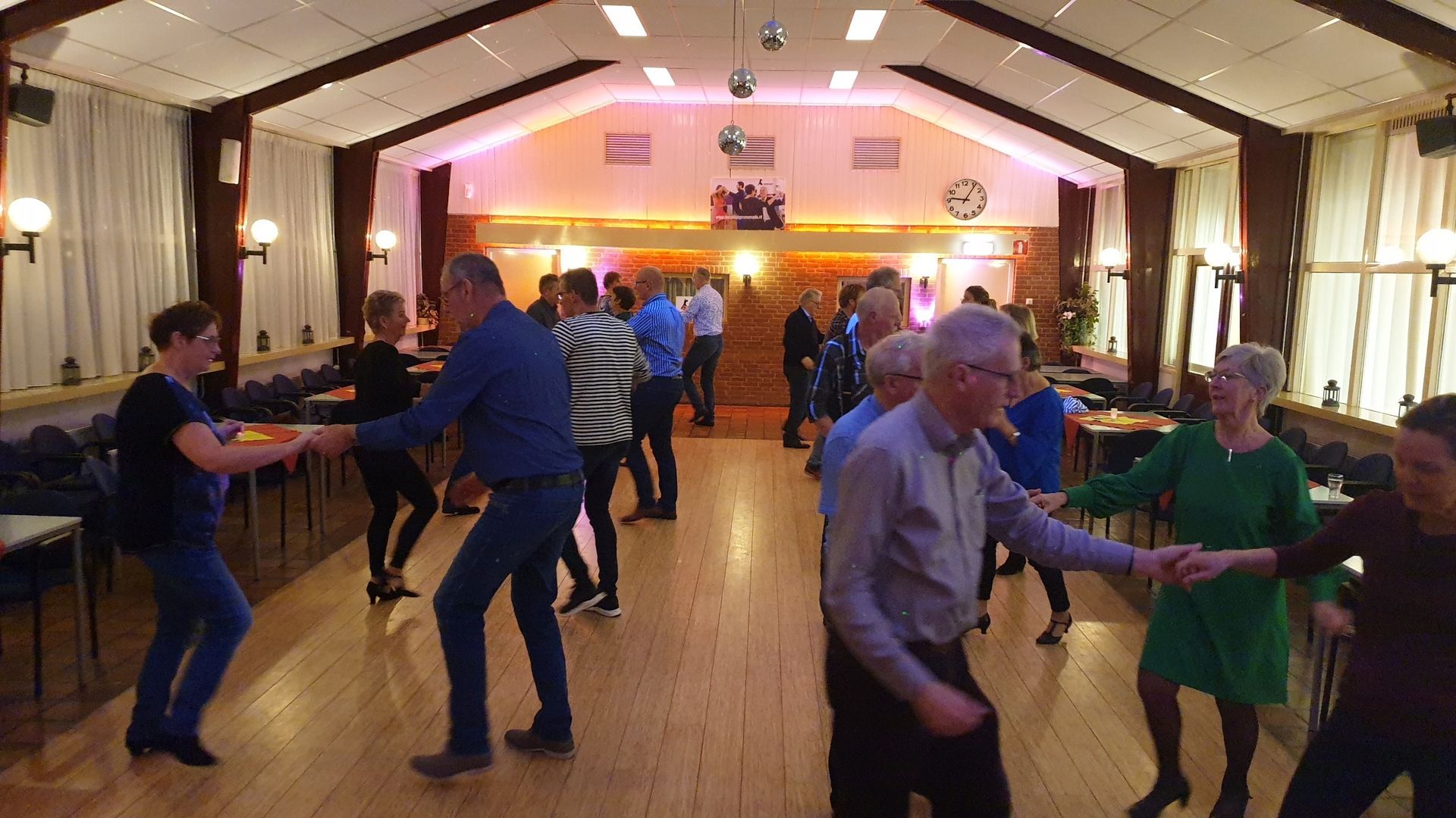 Mensen dansen in een grote zaal met een houten vloer, onder toneelverlichting.