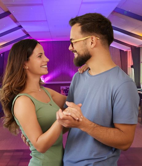Een man en een vrouw dansen heel dicht op elkaar, lachend, in een balzaal met andere paren.