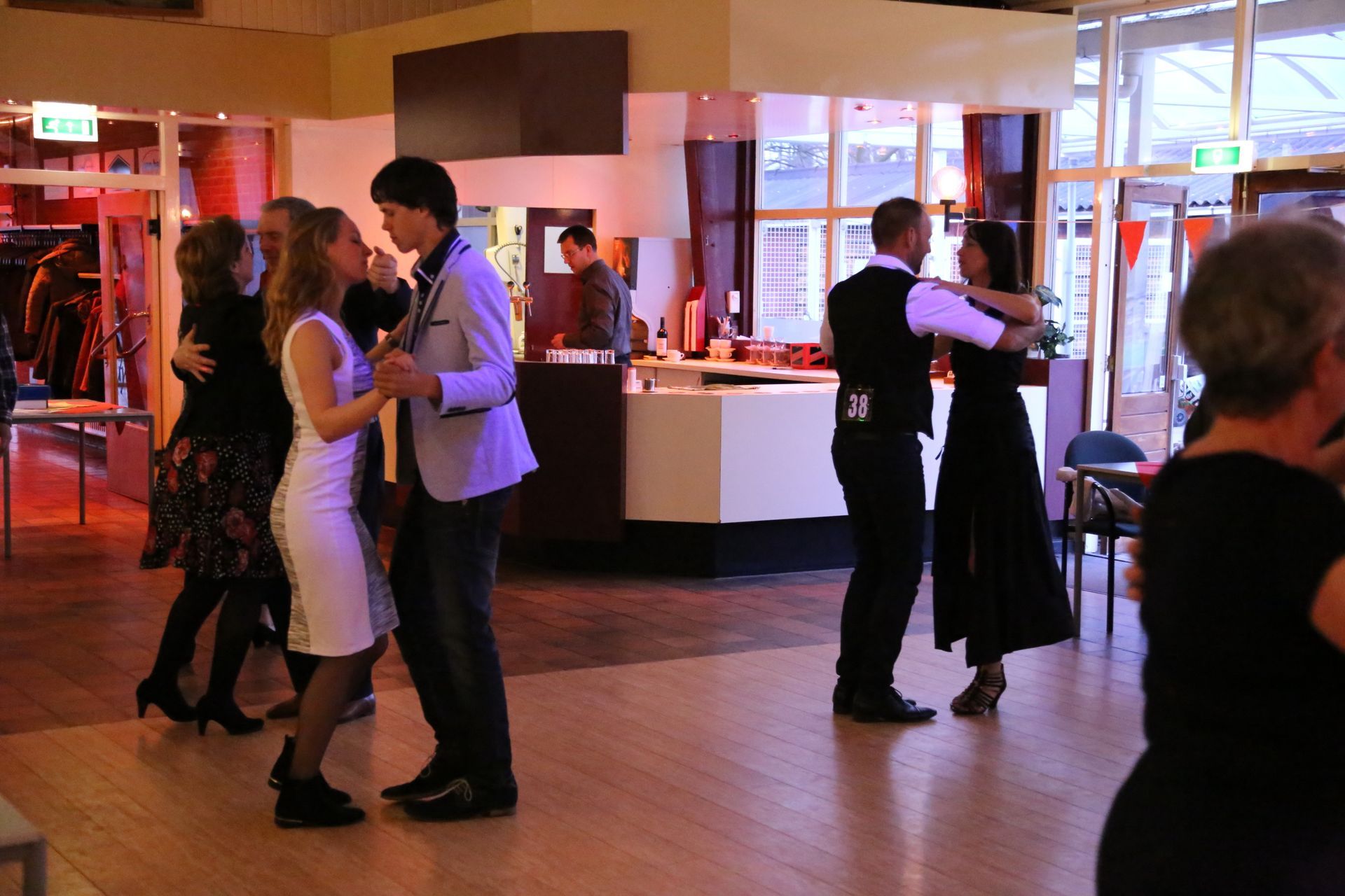 Een lachend stel danst in een dansstudio met anderen. Lichte houten vloer, feestversiering.