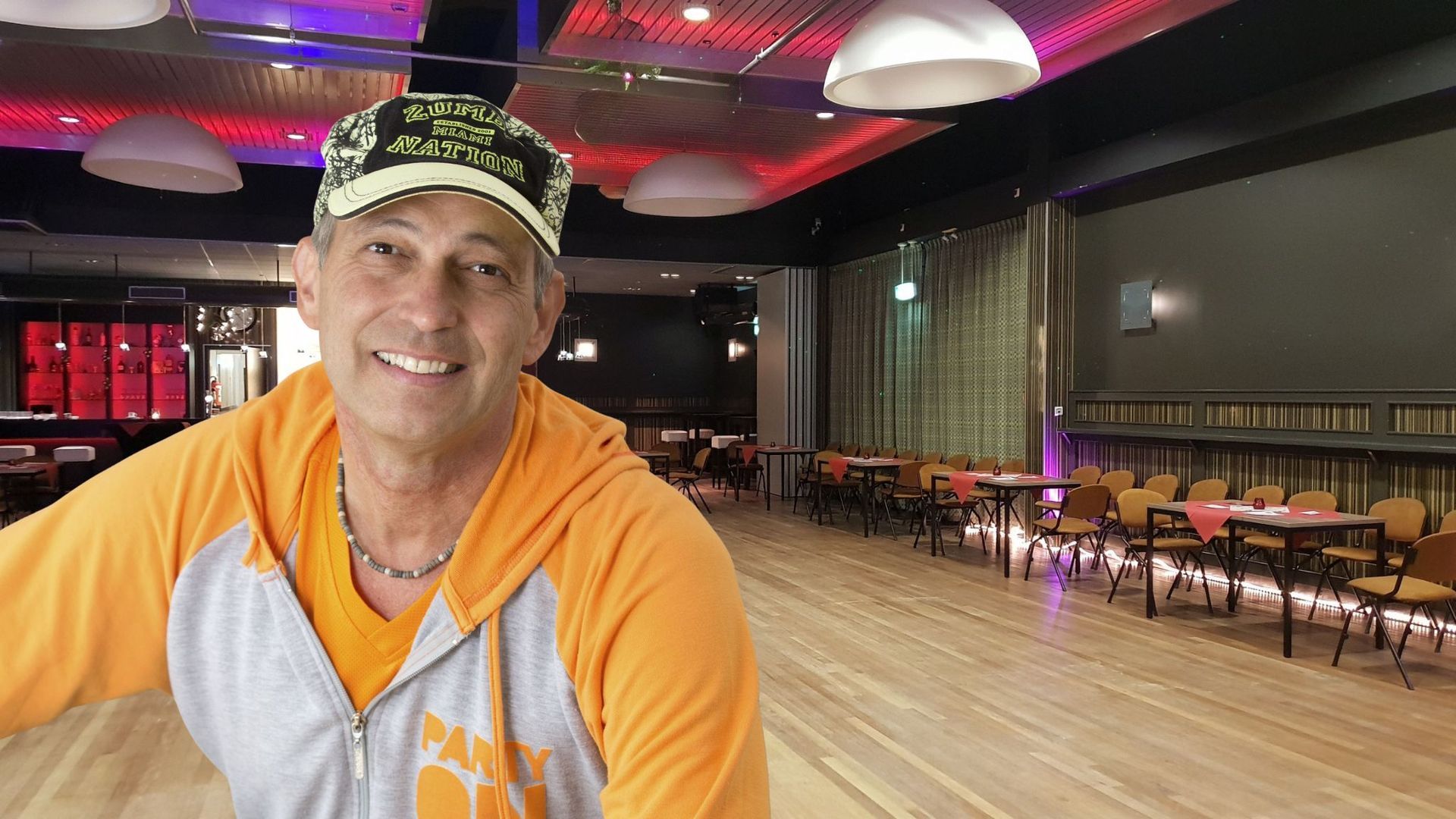 Man in oranje hoodie lacht in een lege balzaal met tafels, stoelen en een podium.