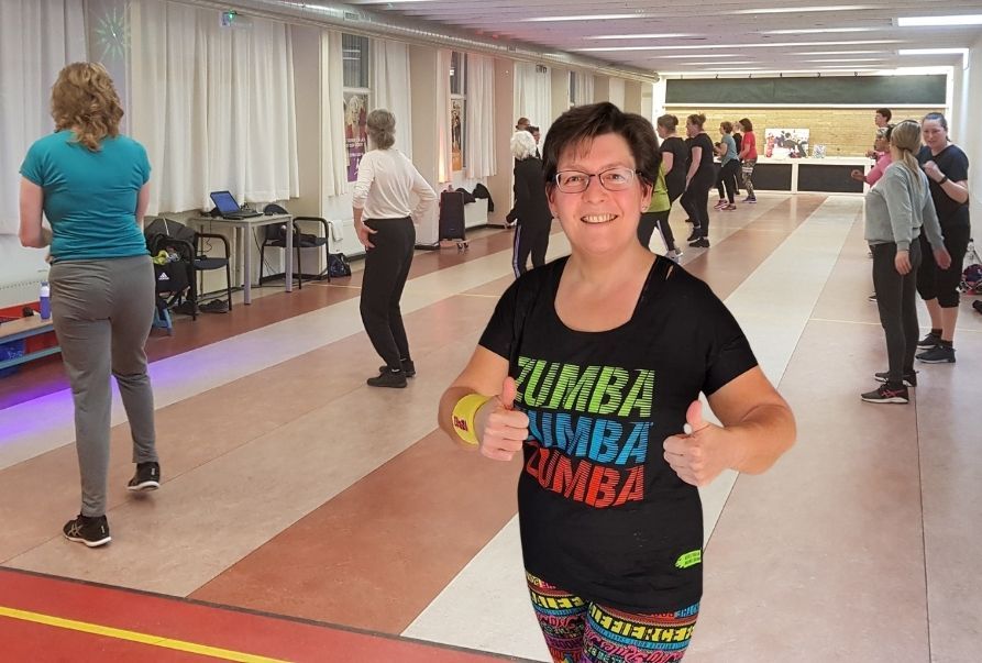 Vrouw in Zumba-shirt lacht en steekt haar duim omhoog tijdens dansles.