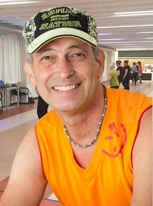Een man in een oranje shirt en pet lacht tijdens een Zumba-les, terwijl anderen op de achtergrond dansen.