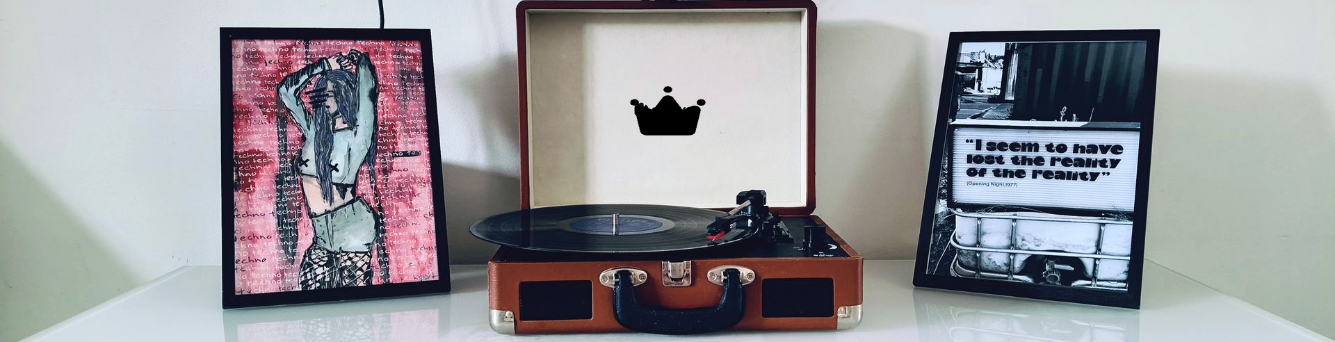 Een platenspeler in de vorm van een koffer in vintage,  tussen twee ingelijste kunstprints op een witte achtergrond.