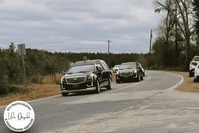 Black vehicles driving on a curved road through a wooded area under a cloudy sky.