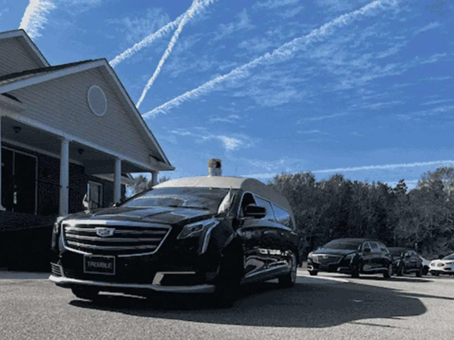 Black hearse and following cars outside a building with blue sky and contrails.