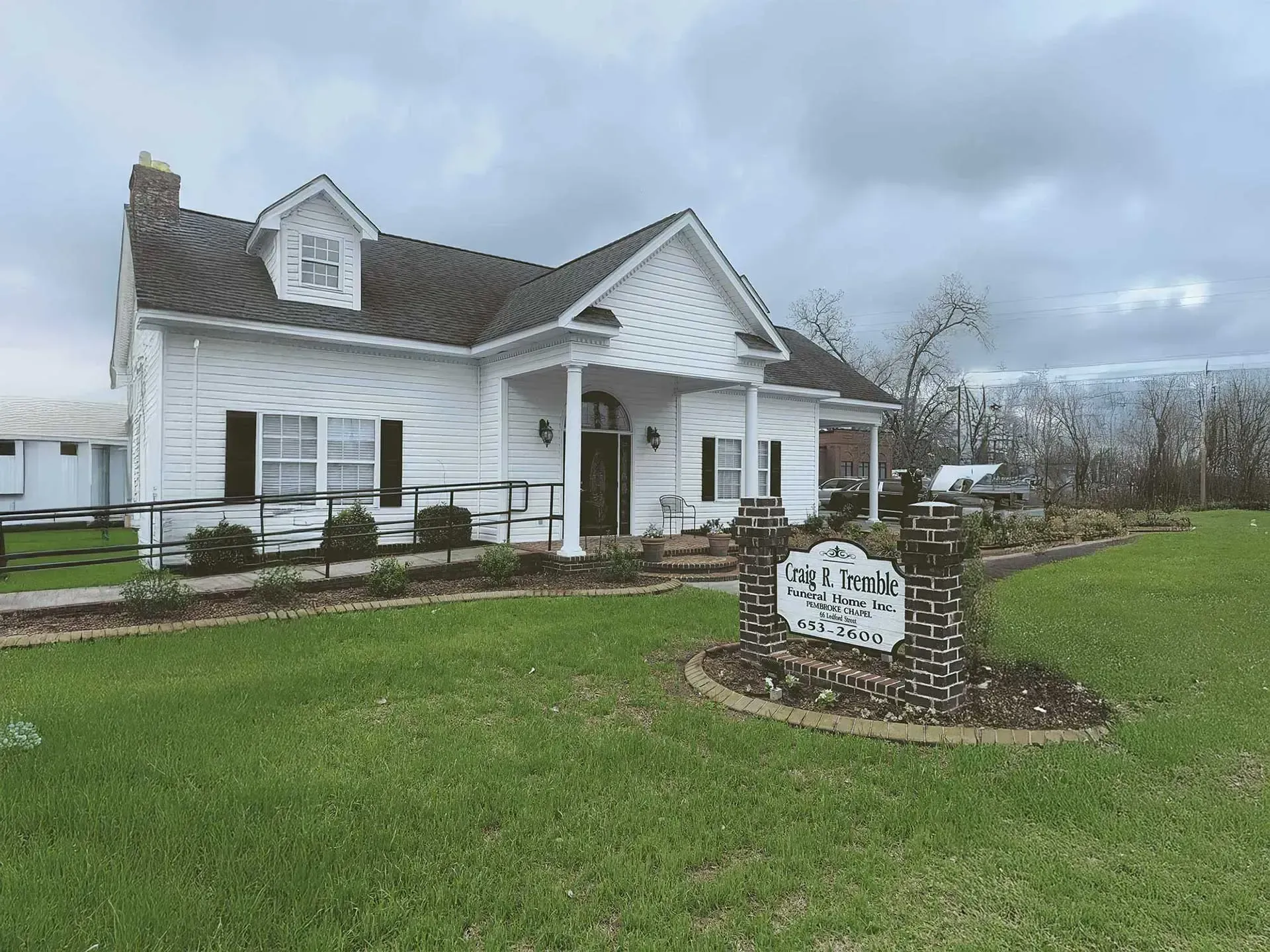 White house with black shutters, sign in front, cloudy sky.