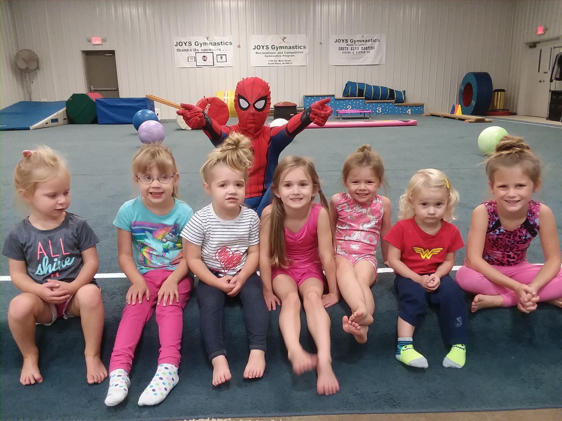 A group of young girls are sitting next to a spiderman mascot.