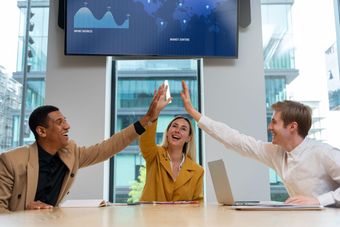 Tres hombres de negocios celebrando con un choque de manos en una oficina, sonriendo frente a una pantalla.