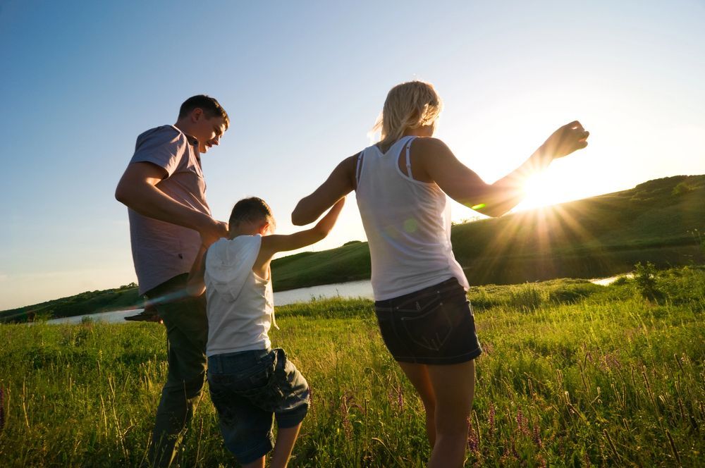 Familia bailando en un campo al atardecer: hombre, mujer, niño, luz solar, pasto verde, agua en el fondo.