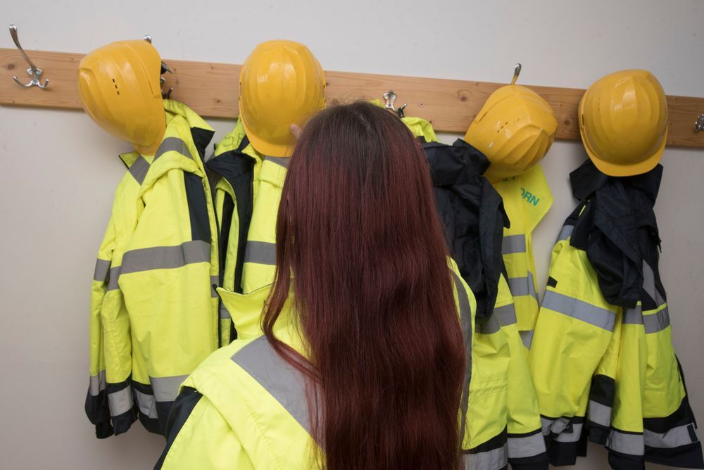 Una mujer con cabello rojo está parada frente a chaquetas de trabajo amarillas y cascos colgados en una pared.