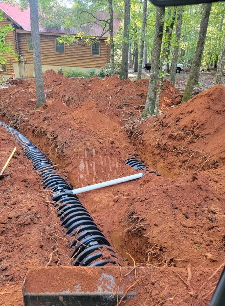 Trenched red clay yard with black corrugated pipe and white PVC near a wooden cabin among trees
