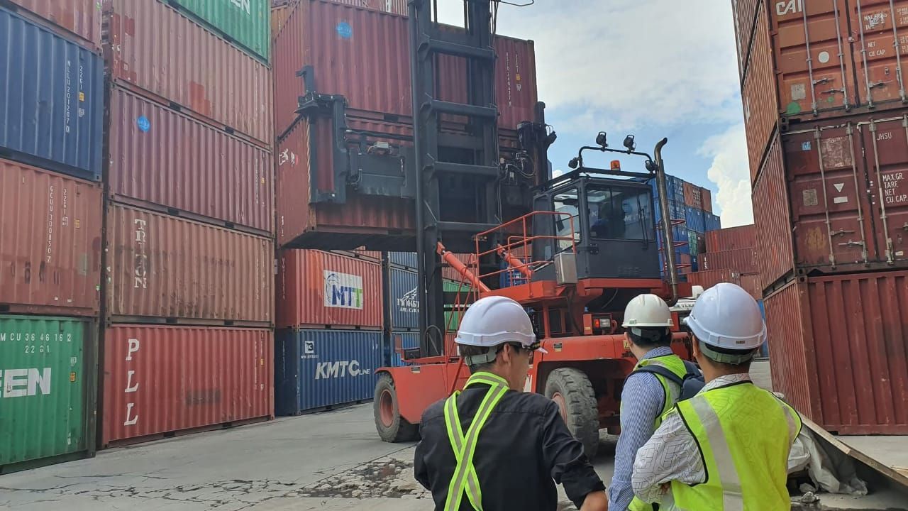 Forklift lifting shipping containers in a port. Workers in hard hats observe. Red, green, and blue containers.
