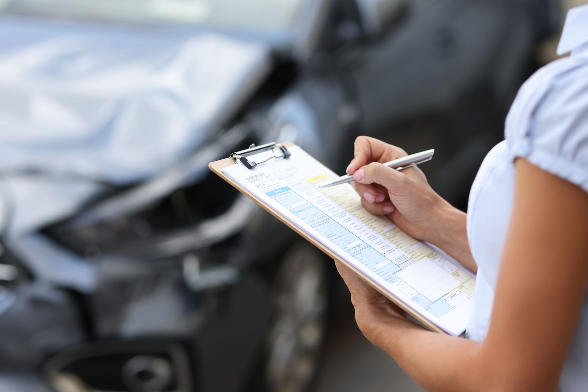 Person writing on a clipboard, assessing damage to a gray car after a collision.