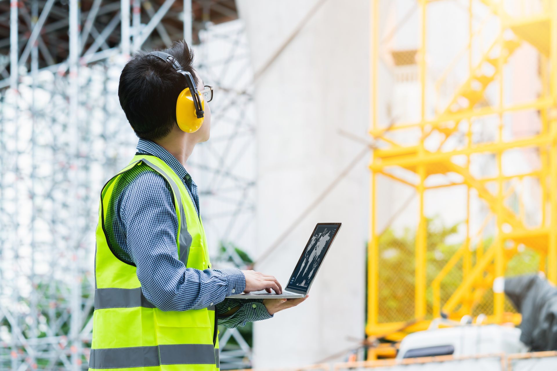 Construction worker in safety vest and earmuffs uses a laptop on a construction site.