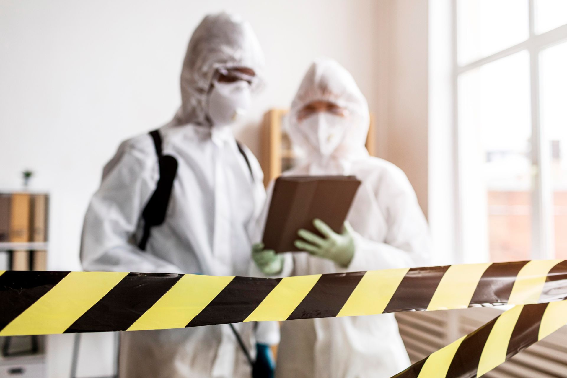 People in protective suits inspect a tablet behind yellow and black caution tape.