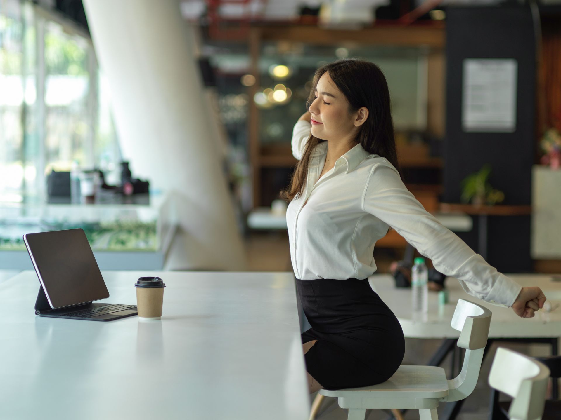 Woman stretching at a desk in an office. She wears a white shirt and black skirt. A tablet and coffee are on the desk.