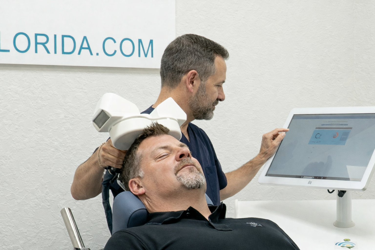 Woman administering TMS therapy to a man seated in a chair, device over his head, near a computer screen.