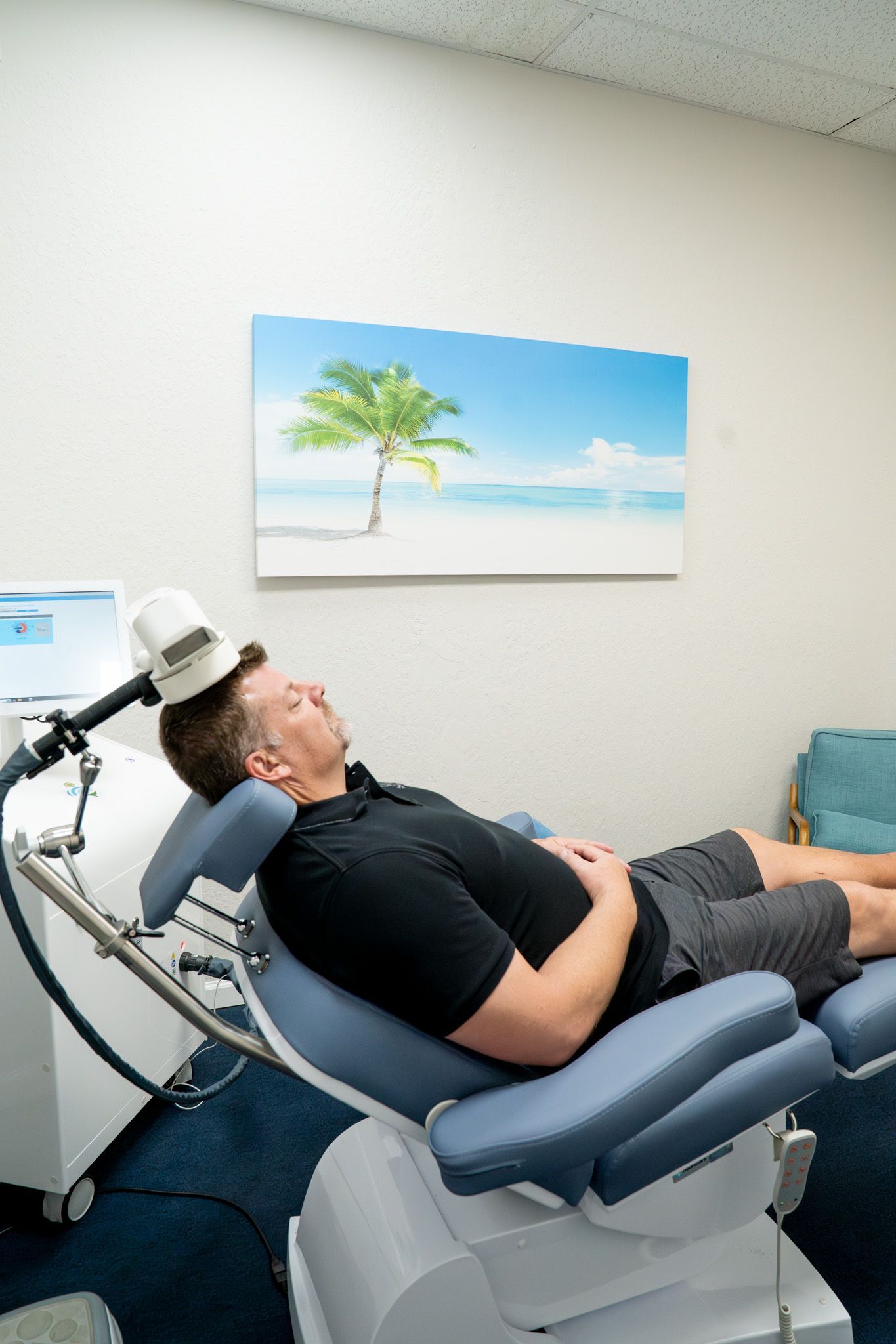 Man receiving TMS therapy, eyes closed, in a medical office, white machine, painting of a beach.