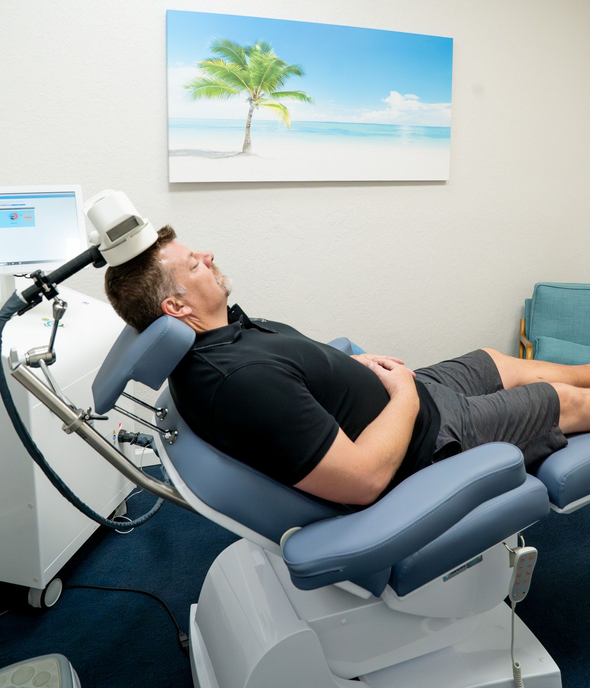 Man receiving medical treatment in a reclined chair; device positioned on his head, beach art in background.