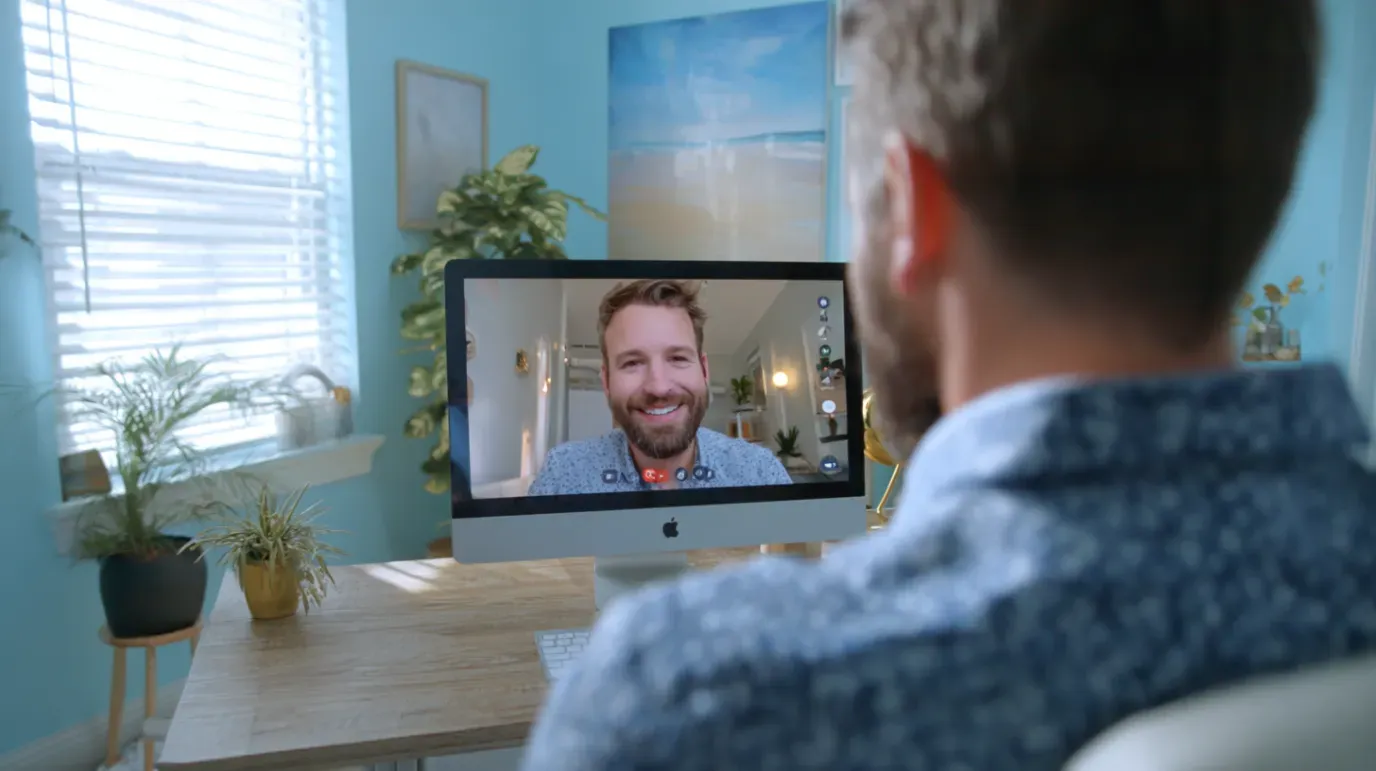 Man on video call with another smiling man on a computer screen. Blue room, window, and desk.