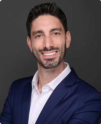 A smiling man with dark hair and a beard, wearing a blue blazer and white shirt, against a gray background.