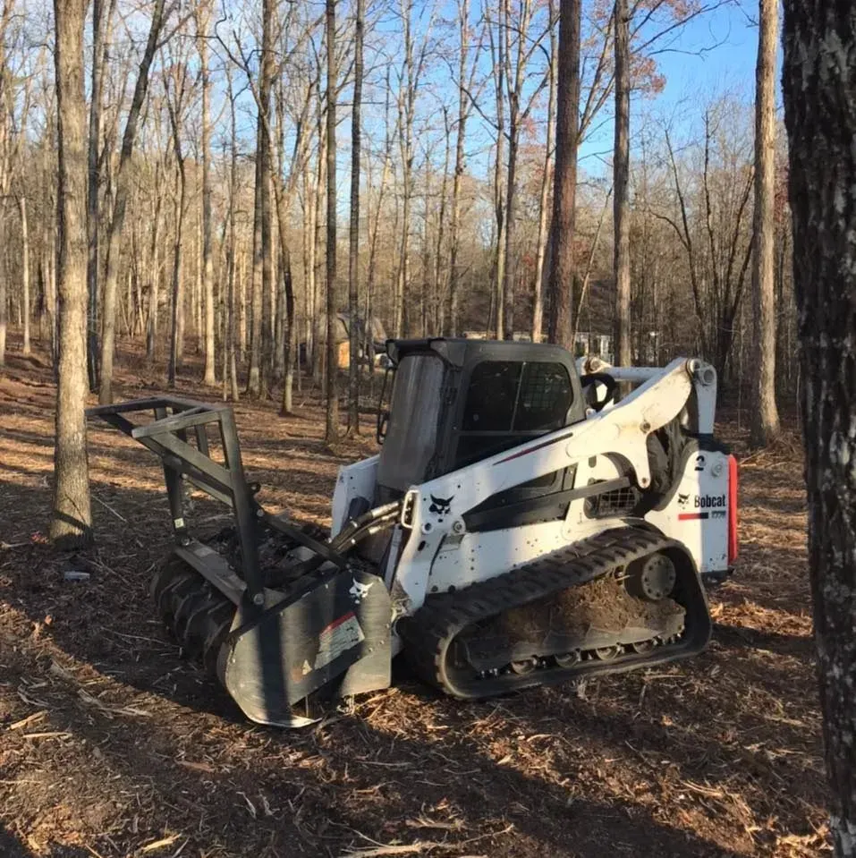 A white and black Bobcat compact track loader with a forestry mulcher attachment sits in a wooded area.