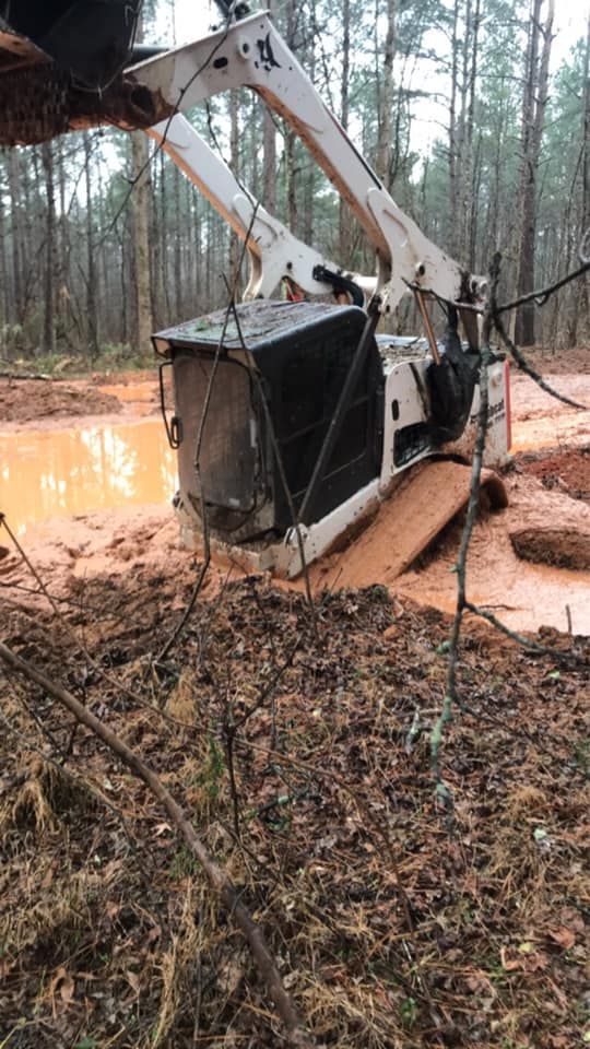 A white skid steer loader stuck deep in orange mud within a forest setting.