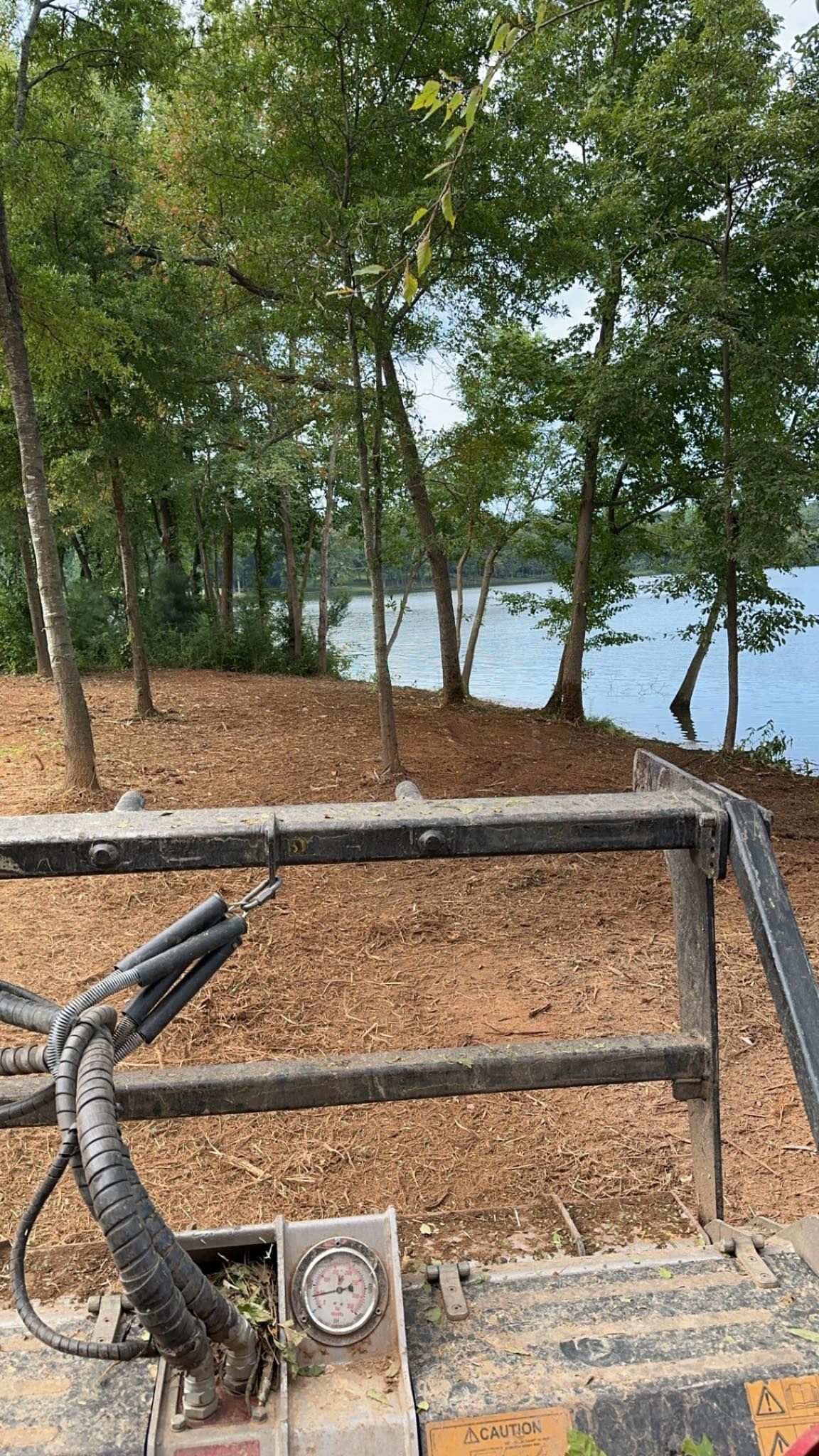 View from the cab of heavy equipment looking over cleared land toward a lake framed by trees.