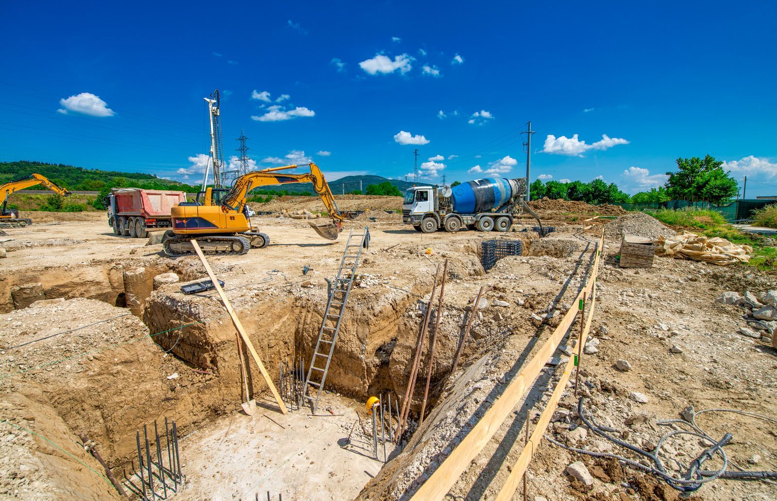 A yellow excavator and a concrete mixer truck working at a sunny outdoor construction site with excavated earth pits.