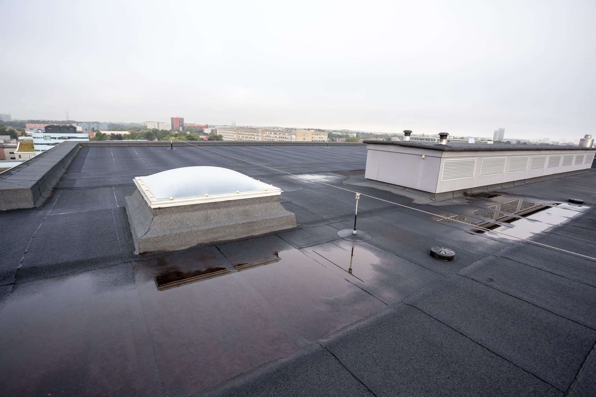 A wide shot of a flat, dark roof featuring a domed skylight, standing water, and a long ventilation structure.