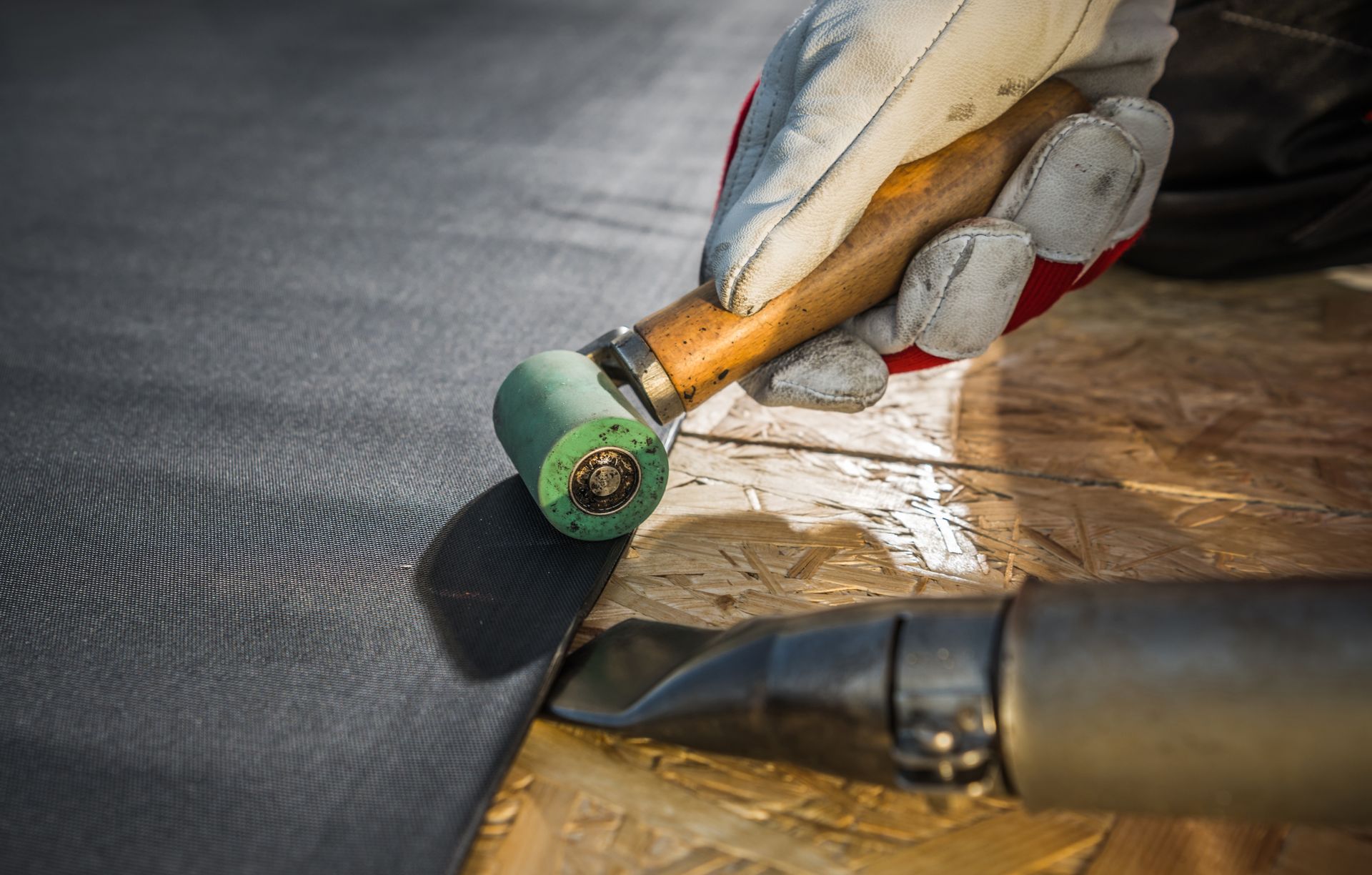 A gloved hand uses a silicone roller and a heat gun to seal a seam on a black rubber roofing membrane.