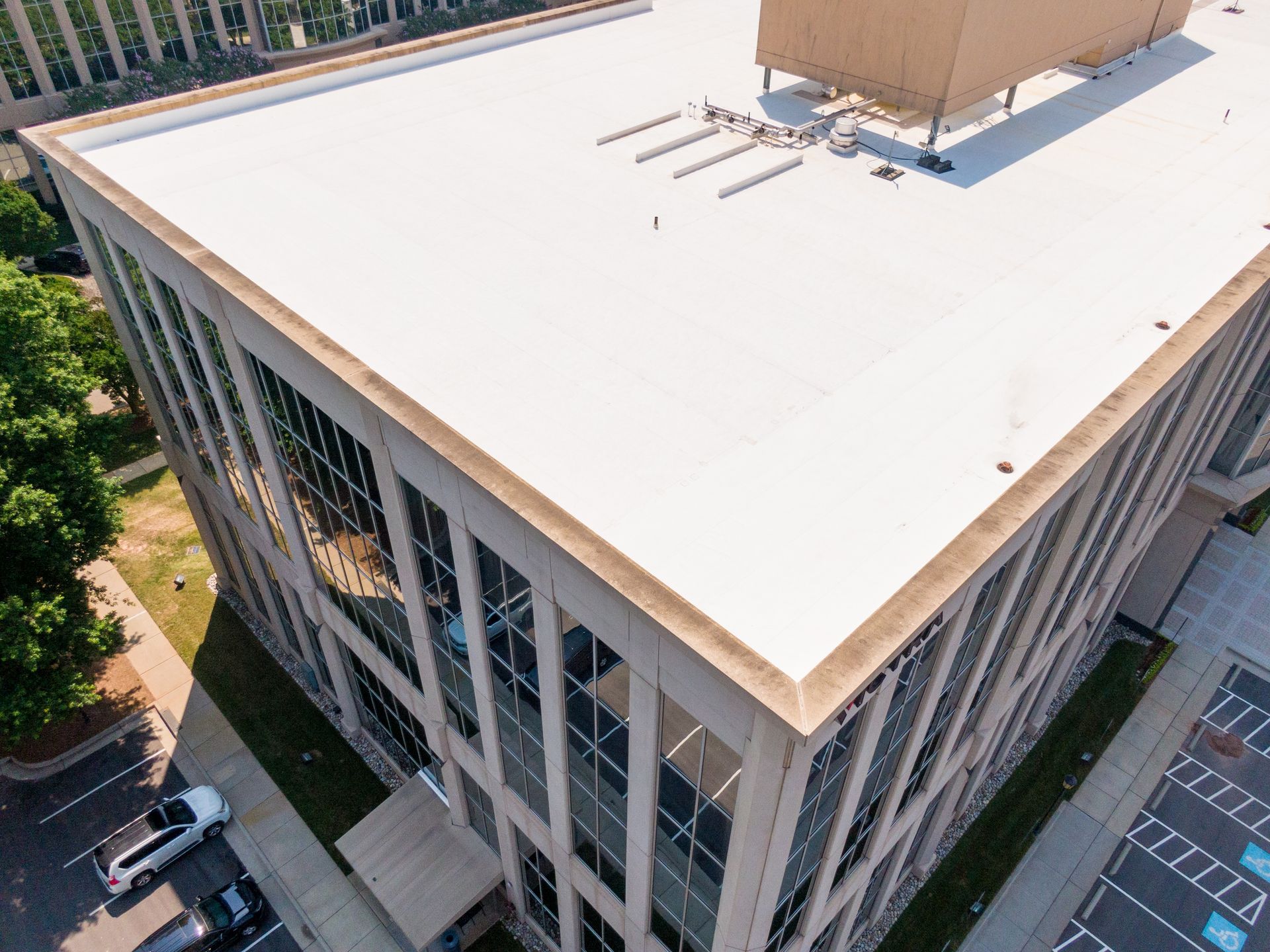 An elevated, angled view of a flat, white-roofed commercial office building with glass windows beside a parking lot.