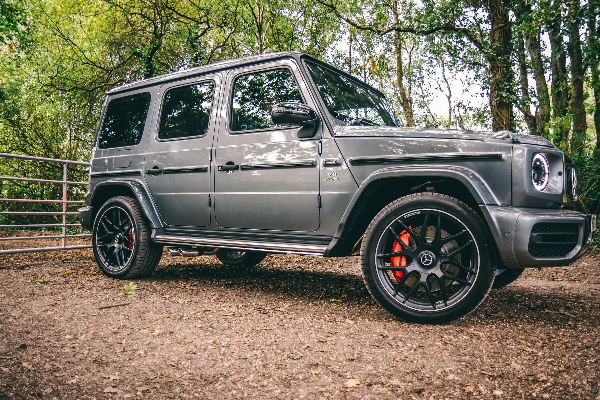 Gray Mercedes-Benz G-Class SUV parked on dirt road, red brake calipers, black wheels, trees in background.