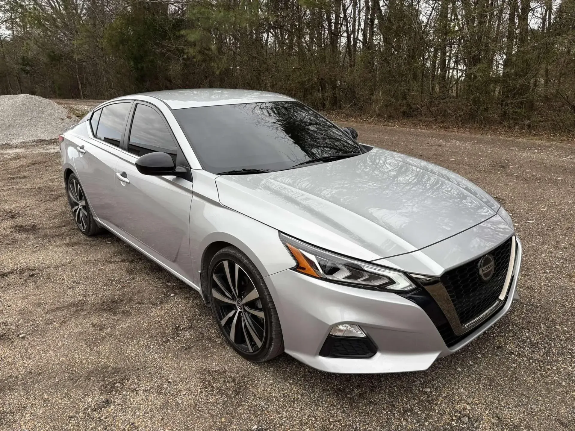 Silver Nissan Altima sedan parked on gravel, with tinted windows and black rims.