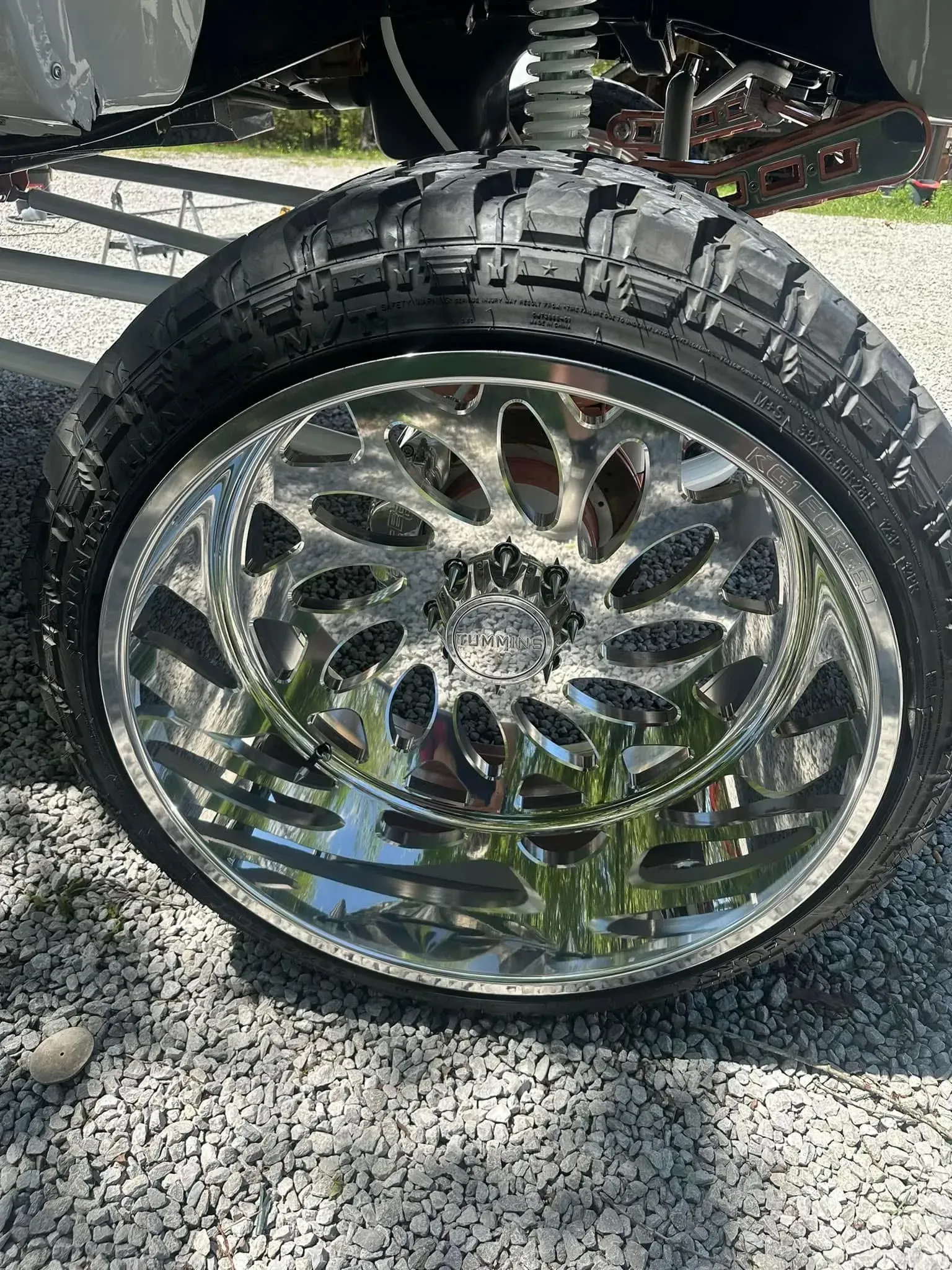 Shiny chrome wheel with black tire on gravel, likely on a vehicle.