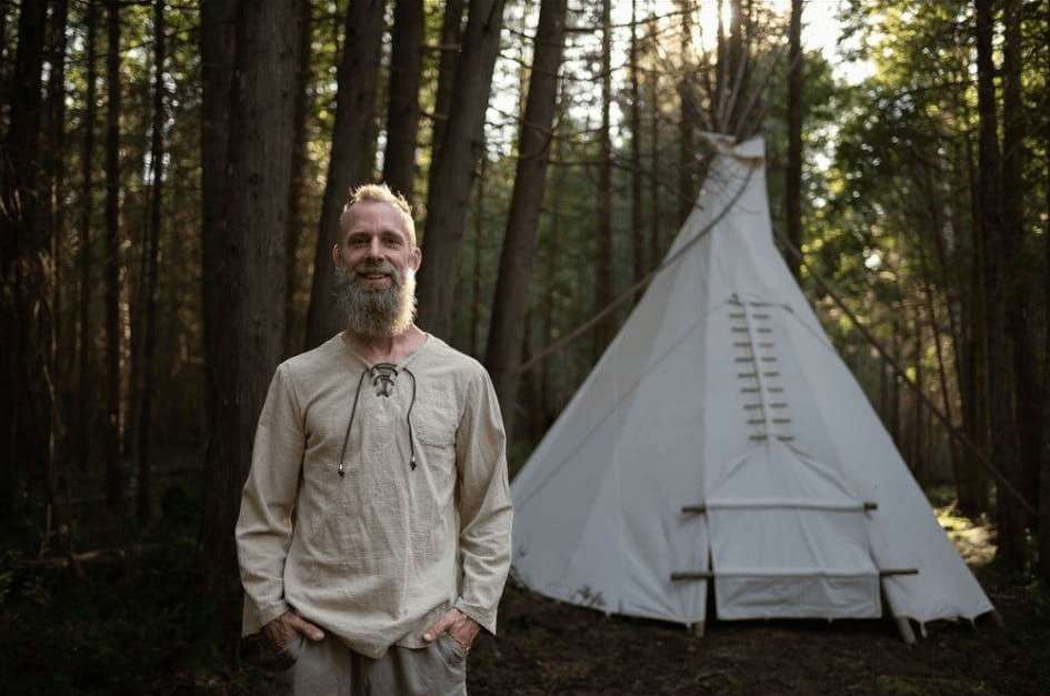 Un homme barbu se tient devant un tipi blanc dans une forêt.