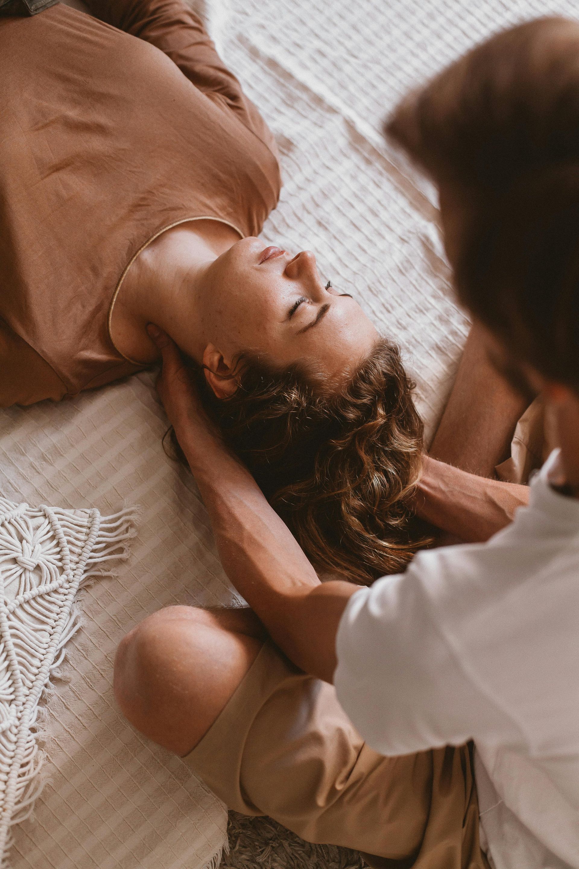 Person lying on a bed while another gently holds their face, in a cozy bedroom setting.