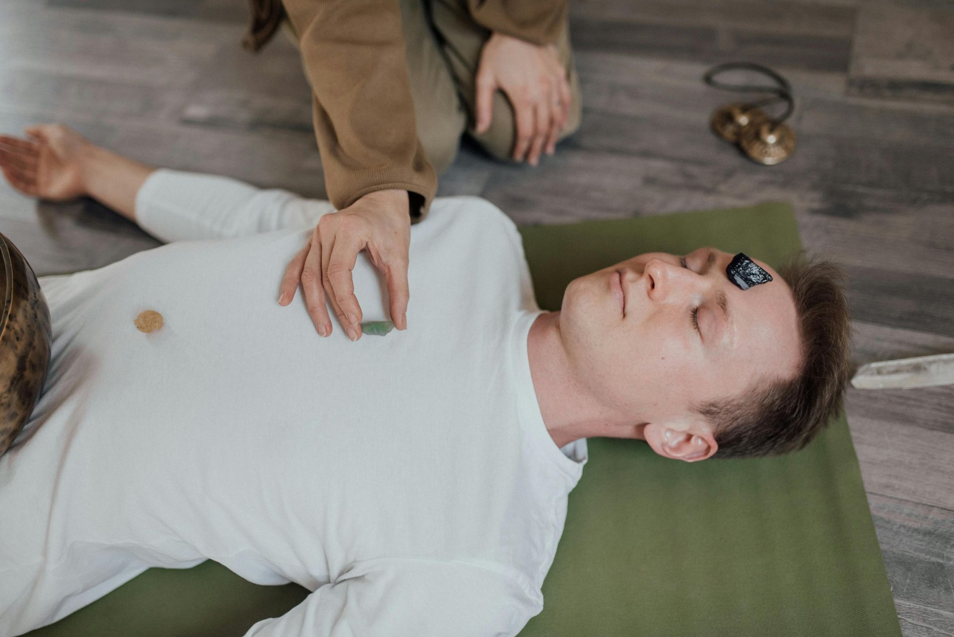 Person lies on a green mat during a healing session while another hand rests on their chest