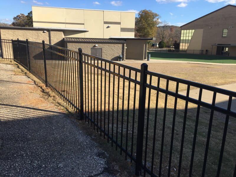 Black metal fence along a gray paved area, with buildings and grassy field in the background.