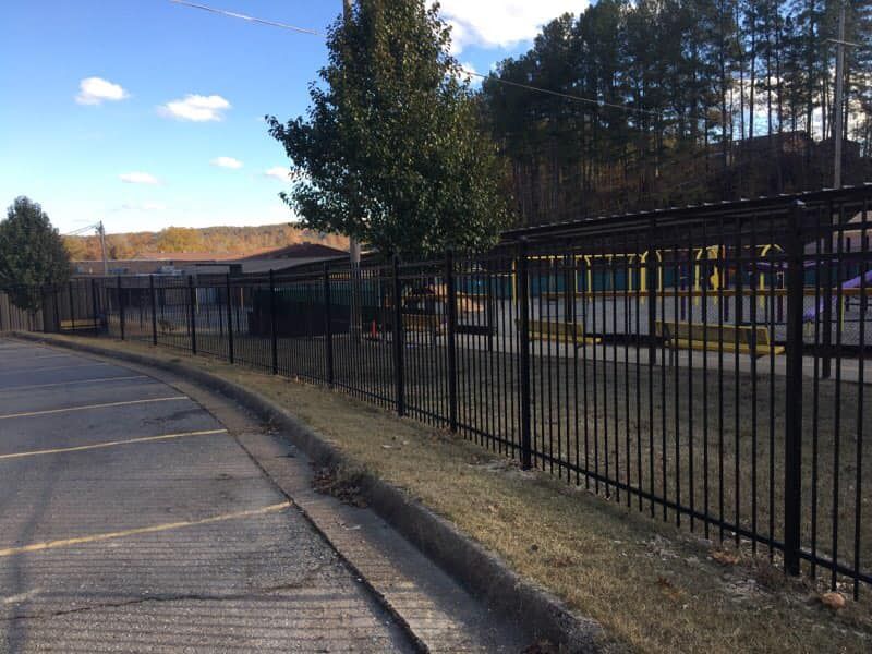 Black metal fence surrounding a building; parking lot and curb in foreground; trees and sky in background.