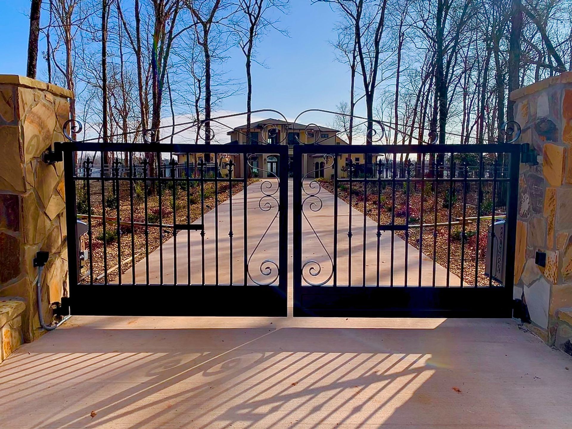 Black iron gates lead to a large home with a long driveway. Stone pillars frame the entrance.