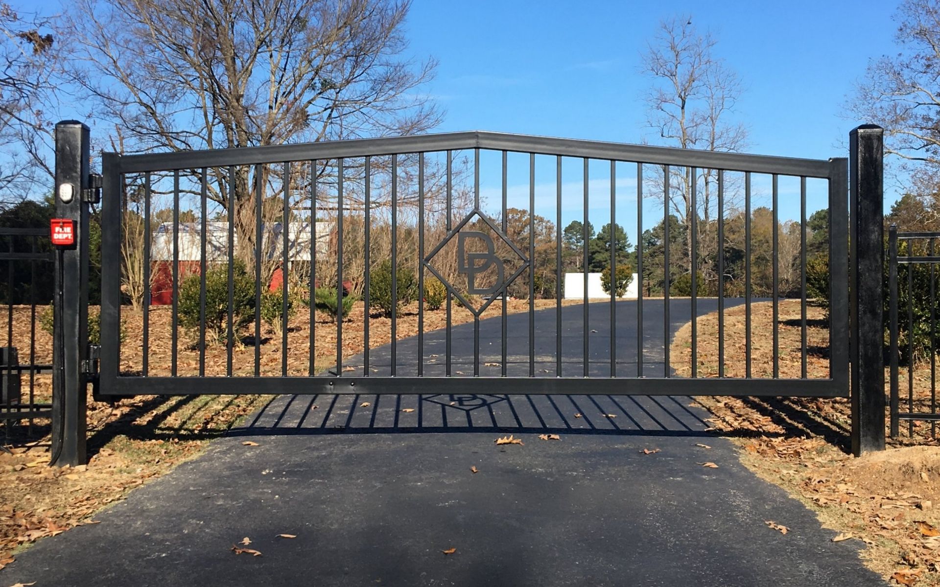 Black metal driveway gate with vertical bars and a diamond design, set in a paved driveway.