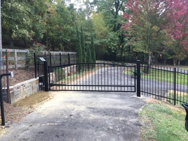 Black metal gate across a driveway. Trees and green grass surround it.